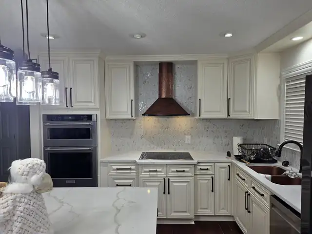 White raised-panel kitchen cabinets with a copper range hood, wall ovens and a marble mosaic backsplash